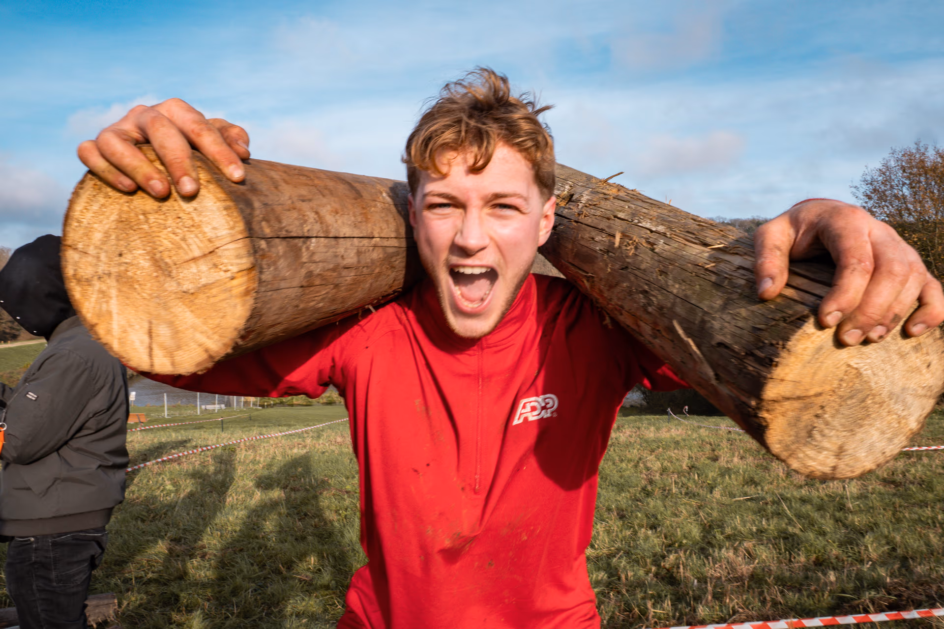 Jeune homme portant des troncs d'arbres sur les épaules lors d'une course d'obstacles, criant vers la caméra de manière énergique
